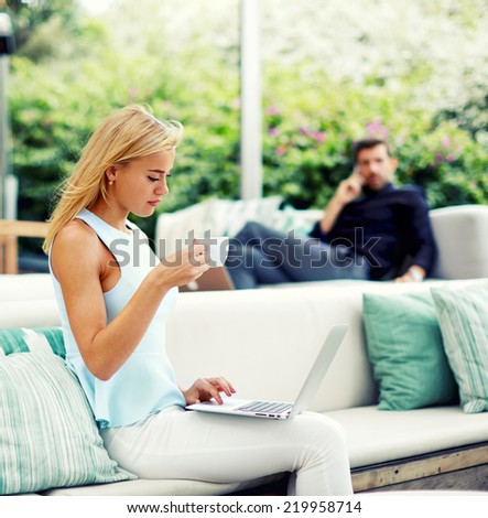 Beautiful blonde hair woman drinking coffee while using computer pc at her work break, handsome rich businessman looking for beautiful woman seated near from him in light modern terrace of lounge bar