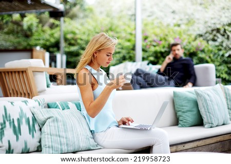 Beautiful blonde hair woman drinking coffee while using computer pc at her work break, handsome rich businessman looking for beautiful woman seated near from him in light modern terrace of lounge bar