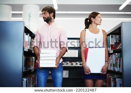 Young international students holding pile of books standing near bookshelf in library, high school students studying hard for exams, students of high school holding stack of books looking away
