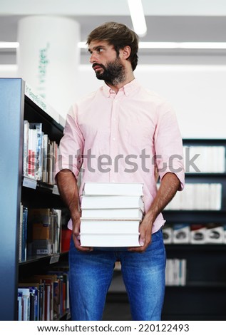Young male student holding pile of books standing near bookshelf in library, attractive brunette student studying hard for exams, learner student of high school holding stack of books and looking away