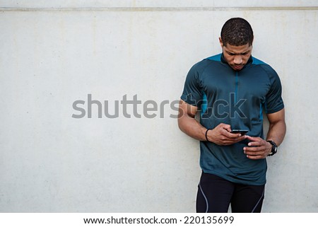 Attractive strong build runner resting after self training standing on white wall background, male runner using mobile phone, dark skinned jogger holding mobile smart phone standing outdoors