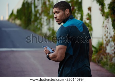 Handsome dark skinned jogger relaxing after workout outdoors, beautiful muscular body man using his mobile phone walking on the road, afro american jogger taking break using  phone standing outdoors