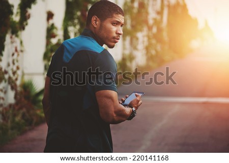 Male sportsman walking on the road to sunset holding phone in the hands, athletic build male runner holding mobile phone, attractive jogger using smart phone after self training outdoors