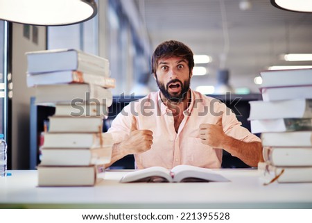 Young undergraduate ready for his final exam, surprise emotion face of student sitting at the library desk with two pile of books,student with two thumbs up make positive facial expressions