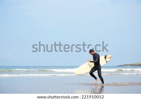 Young surfer man carrying his surfing board running along the ocean to waves, guy with surfboard running touching waves, professional surfer man dressed in wetsuit ready to surfing runs to the waves