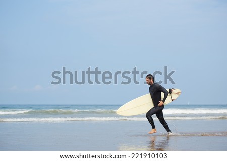 Young attractive surfer runs with surfboard along the shore, cold season surfing at ocean beach, professional surfer man dressed in wetsuit ready to surfing runs to the waves