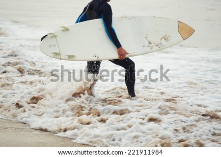 Man surfer with surfboard, surfer man carrying his surfing board, surfer in wetsuit holding a surfboard, surfer walking on the beach touching waves, surfer going to the water