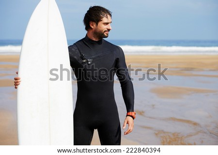 Muscular build surfer holding his surfing board standing on ocean beach with big waves in background, young surfer standing on a beach with his surfboard and looking along the shore, healthy lifestyle