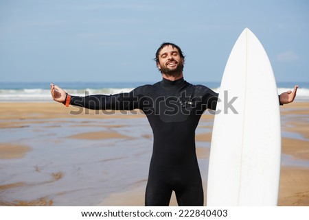 Feeling good and happy with healthy lifestyle, attractive surfer man enjoying perfect sunny day standing on the beach and holding with one hand his surfboard, young surfer feeling so happy