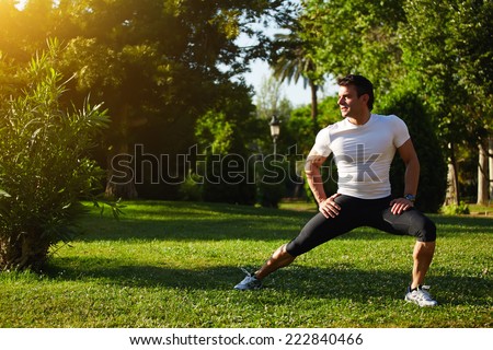 Fit man doing stretching exercises outdoors, young male sportsman stretching and preparing to run, attractive adult runner stretching the legs standing on grass in the park, sports fitness concept