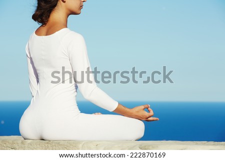 Yoga meditation on high altitude with blue sea on background, woman meditating yoga enjoying sunny evening, meditating woman, beautiful woman in white clothes seated in yoga pose outdoors