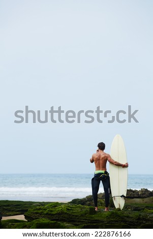 Professional surfer man in black wetsuit standing on the rocks covered with moss enjoying beautiful view of ocean, surfer holding his favorite surfboard looking for the ocean waiting big waves