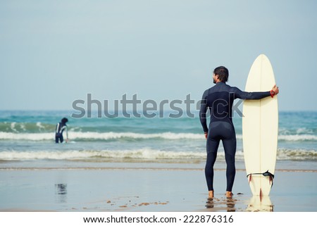Muscular build surfer holding surfboard while standing on the beach looking at ocean to find the perfect spot to go surfing waves, professional surfer waiting waves on the ocean beach, filtered image