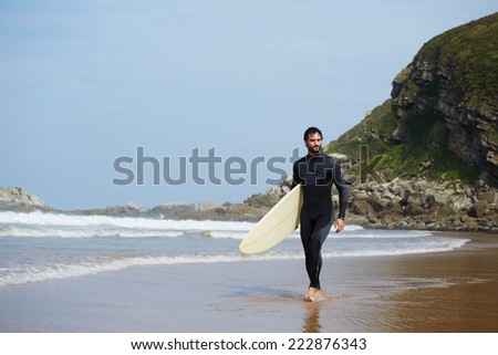 Handsome surfer walks carrying surfboard with big mountain rock on background, perfect sunny day for surfing on big waves, young handsome surfer man walking with surfboard on the beach