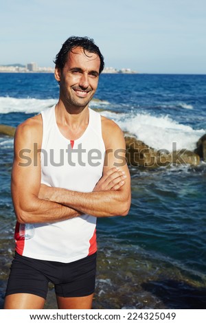 Fit male jogger standing on seaside while resting after intensive fitness training, fit man in bright sportswear smiling taking a break, portrait of smiling runner resting after intensive run outdoors