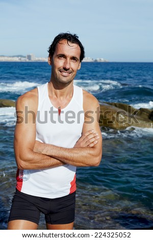 Portrait of smiling runner standing on rocks at sunny summer day, attractive male jogger standing on seaside while resting after intensive fitness training, fit man in bright sportswear taking break