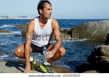Male fit runner resting while sitting on rocks with blue sea on background, attractive runner holding bottle of water taking a break after workout, man resting after doing sports outdoors