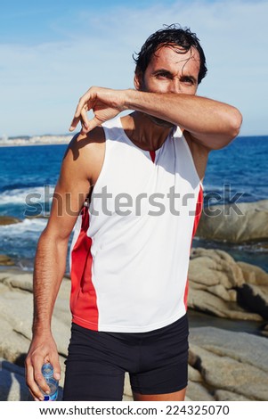 Tired athlete rubs the face after drink cold water, fit man in bright sportswear taking break standing on sea rocks, exhausted runner after workout resting while standing on the beach
