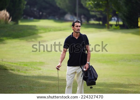 Confident rich man at golf game standing on beautiful green golf course holding bag and golf ball, handsome rich man at his favorite hobby, male golf player at the course with a club sack
