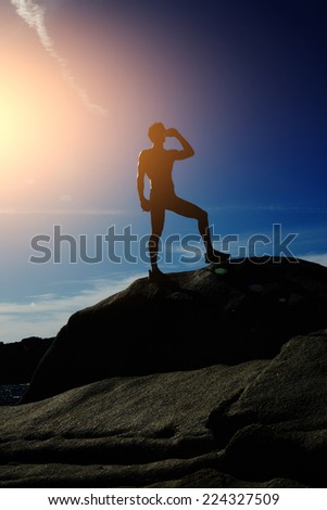 Silhouette of young man drinking water after jogging standing on the rock, athlete refreshing with bottle of water after running