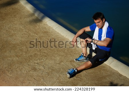 Athletic build jogger sitting in the park taking a break after run, athlete runner with towel on the neck resting after jogging, athlete sportsman checking the time on his wear watch