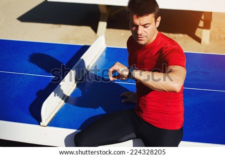 Muscular build jogger looking the time on his watch taking a break after run, athlete runner seated on the tennis table resting after jogging, athlete sportsman checking the time on his wear watch