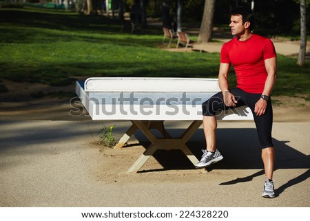 Attractive sportsman sitting on the tennis table resting after intensive evening run, beautiful fit man with perfect muscular body taking break in beautiful park at sunny summer evening, sport concept