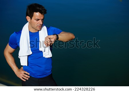 Muscular build jogger looking the time on his watch taking a break after run, athlete runner with towel on the neck resting after jogging, athlete sportsman checking the time on his wear watch