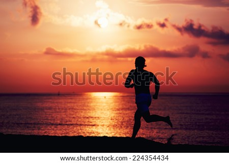 Silhouette of muscular build athlete running fast a log the beach, runner in action jogging against colorful sunrise over the sea