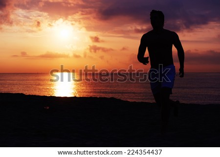 Dark silhouette of running man towards colorful sunrise at early morning, male runner with muscular body at fitness training jogging on the beach