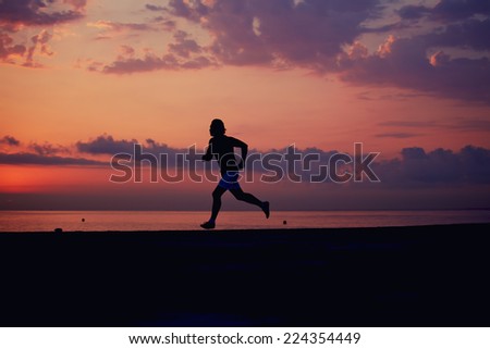 Perfect and beautiful silhouette of sporty fit man running on the beach, morning fitness training outdoors, young jogger running fast at colorful sunrise