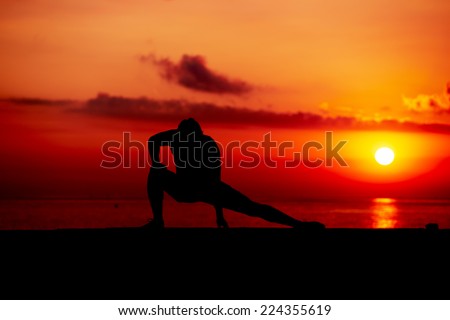 Silhouette of young man at cross-training during colorful sunrise on the beach, athletic runner with muscular body doing stretching legs exercise outdoors, fitness and healthy lifestyle concept