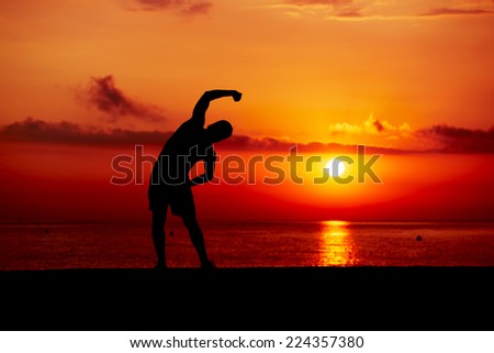 Silhouette of handsome muscular man doing exercise against colorful sunrise sky, sportsman doing stretching exercise standing on the beach