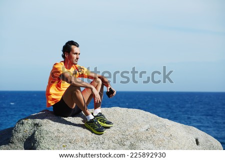 Attractive sportsman in bright sportswear listen music enjoying sunny summer day on the beach, fit man listening to music on his mobile smart phone while resting after run outdoors, enjoying music