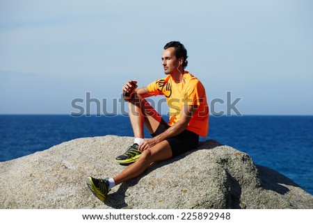 Relaxed fit man listening to music sitting on sea rocks outdoors, fit man in sportswear taking break sitting on rocks with sea horizon on background, rest and relax after the training, sport lifestyle