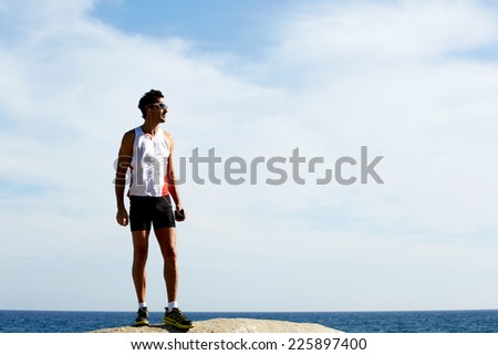 Exhausted fit runner after workout resting on seaside, sportsman listening to music with headphones, athlete standing on sea rocks at sunny summer day while resting after intensive training outdoors