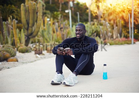 Handsome dark skinned jogger relaxing after workout outdoors, beautiful sportsman using his mobile phone sitting on the road, afro american jogger taking break using phone seated outdoors