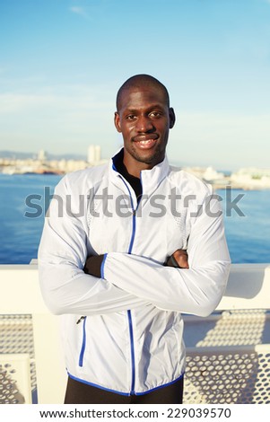 Portrait of smiling man resting after workout outdoors, fit black man with arms crossed standing on modern bridge over the sea near big city, smiling sportsman resting after training outdoors