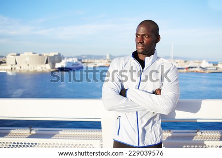 Sportsman in white wind breaker taking break with crossed hands standing on sea background, black fit man resting after workout outdoors,tired to run dark skinned runner resting after fitness training