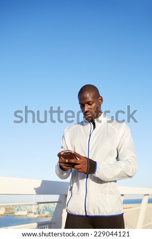 Male runner in white windbreaker resting after run while typing something on his mobile smart phone, black man holding smart phone resting after fitness training outdoors, people using technology