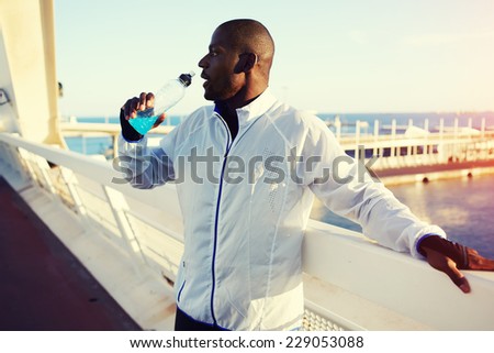 Attractive fit man drinking water after intensive workout training outdoors, black man in windbreaker resting after run