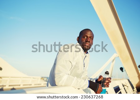Handsome fit man dressed in wind breaker taking a break leaning on bridge fence, male runner holding energy drink looking away while resting after run, evening fitness training outdoors