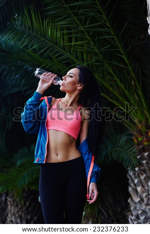 Charming fit woman with sexy abdominal refreshing with water after run, attractive healthy woman with beautiful body dink water after workout outdoors, female jogger taking a break after jogging