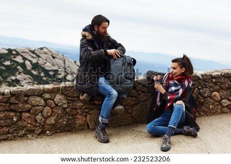 Young hikers resting after long hike way sitting on mountain hill, attractive hiker man with beard searching something in his rucksack, pretty hiker girl holding bottle of water in the hands resting