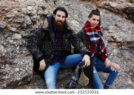 Man and woman hiker seated on rock cliff looking to the camera resting after hike, hipster man with beard and charming hipster girl in mountain landscape nature, mountain walk