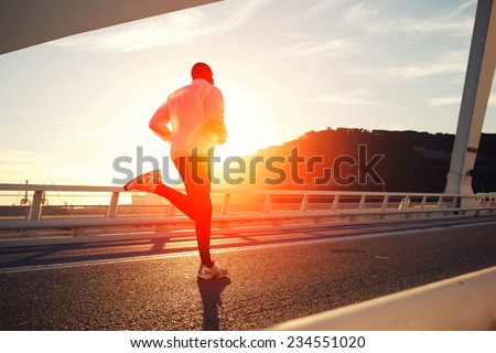 Attractive fit man running fast along big modern bridge at sunset light, black man doing workout outdoors, silhouette runner in windbreaker jogging over bridge road with amazing sunset on background