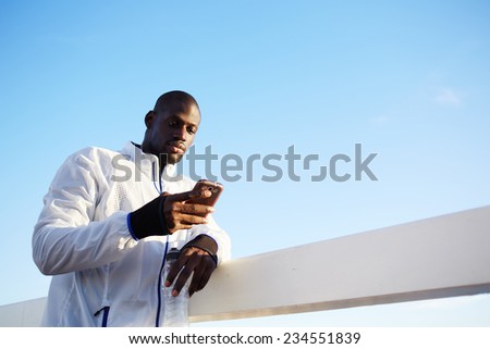Male runner in white windbreaker typing something on his mobile smart phone standing at sunset on beautiful road, black man holding smart phone resting typing something,fit man with mobile smart phone