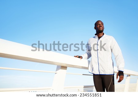Young sportsman in white wind breaker taking break standing on sea background, black fit man resting after workout outdoors, soft evening light, tired to run jogger resting