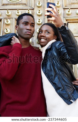 Outdoors portrait of smiling couple taking photos with a smart phone, cheerful trendy couple taking self-portrait picture, best friends fooling around making self portrait with digital camera on phone