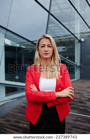 Caucasian businesswoman with arms crossed standing against her office building looking to the camera, blond hair executive business woman dressed in red jacket standing near skyscraper office building